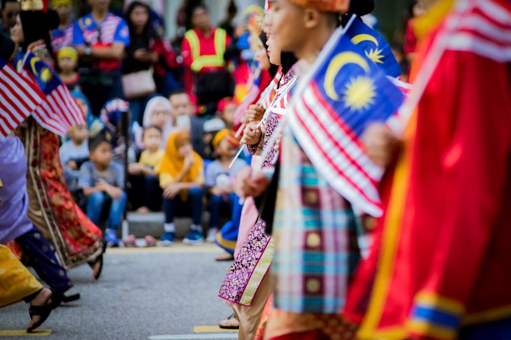 Colorful Malaysian cultural parade with people waving flags and traditional attire, celebrating unity and diversity, emphasizing ARCO Alliance Org's community engagement and multicultural values.